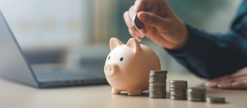 Hand dropping coin into piggy bank beside stacked coins and laptop