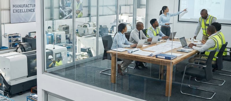 Manufacturing team reviewing production plans in a glass meeting room above the factory floor
