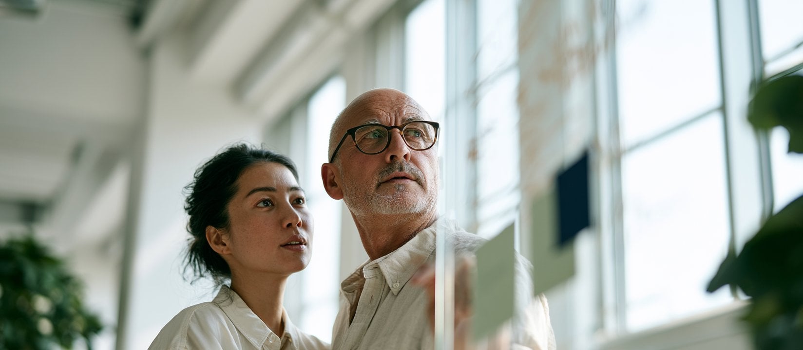 Two colleagues review notes on a glass board in a sunlit office