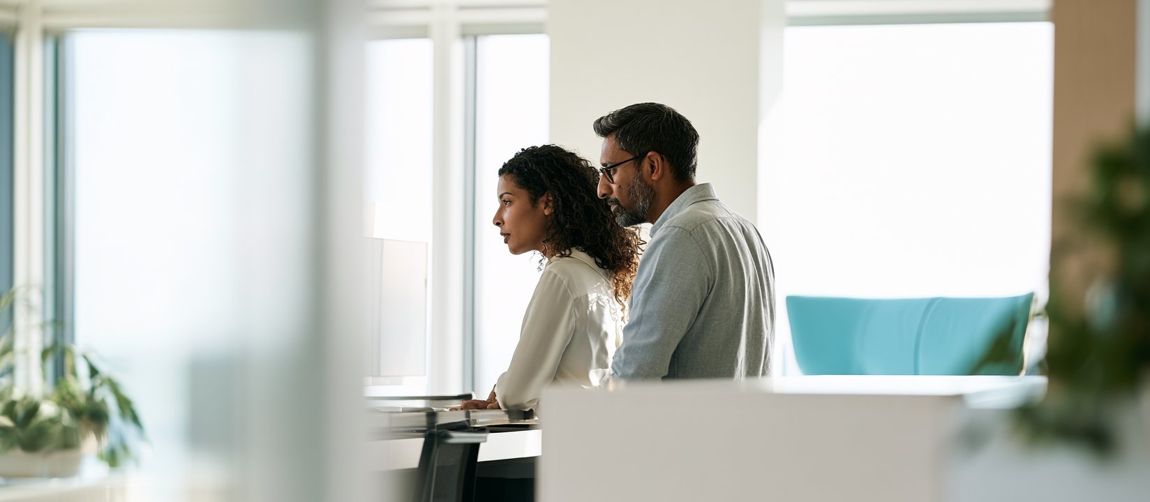 Two coworkers review information on a desktop screen in a bright office