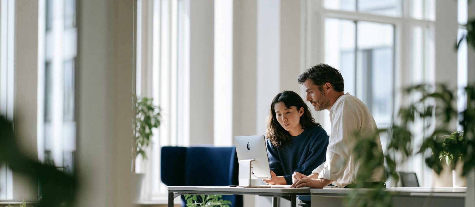 Two coworkers review work together at a computer in a sunlit office