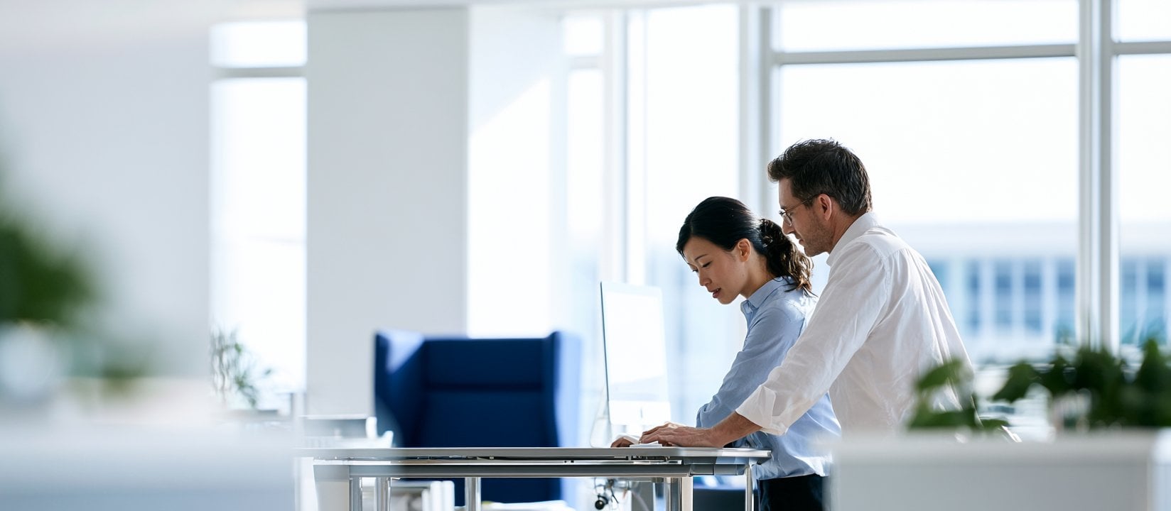 Two coworkers stand at a desk reviewing work on a desktop monitor