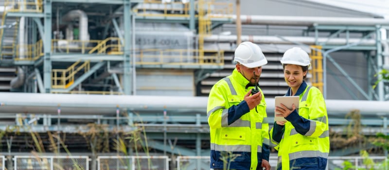 Two workers review a tablet and radio outside an industrial plant