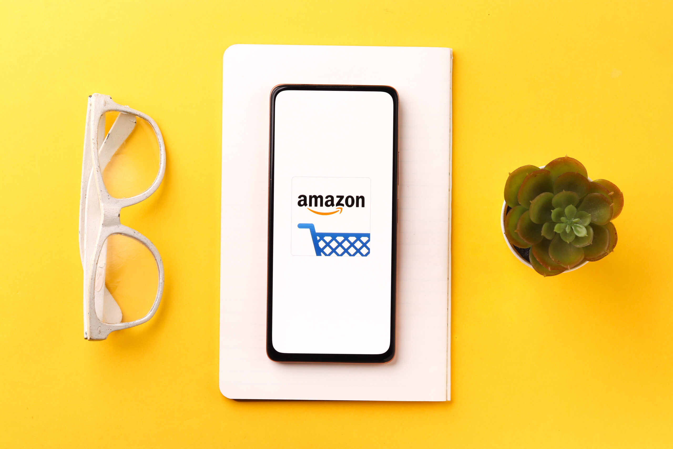 Amazon-Preissetzungsstrategie-Smartphone-auf-gelbem-Tisch-mit-Brille-und-Pflanze